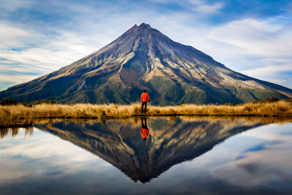 Man stands facing a mountain, reflected in a clear lake