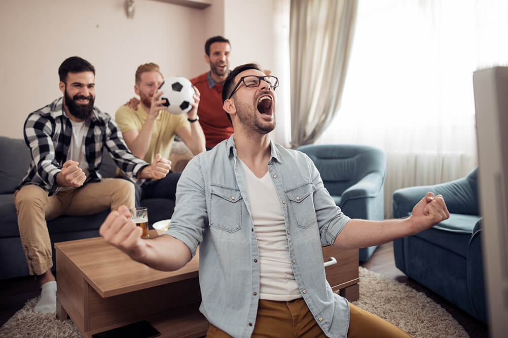 Four men celebrate their soccer team scoring as they watch on TV in the front room.