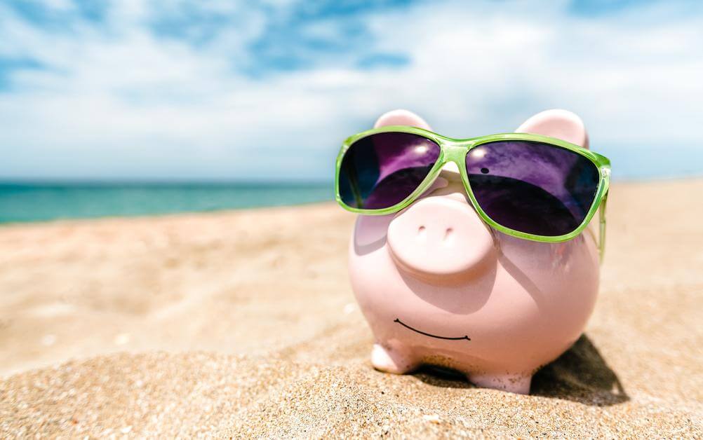 A piggy bank wearing sunglasses on the beach which wont be needed to play the cheapest lottos