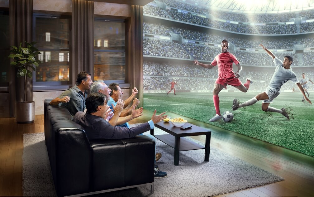 Live Betting Explained A group of friends watching soccer on a huge TV, celebrating as the live betting in-play bet wins.
